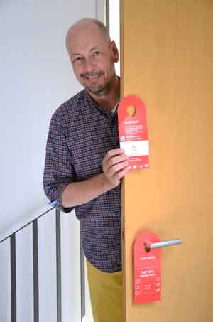 Ein Mann steht halb verdeckt hinter einer Tür und hält einen rotes Türschild in der Hand.