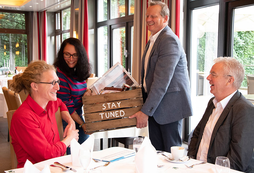 Daniel Gewand und Johannes Arntz sitzen links und rechts am Tisch. Rosalia Rodrigues und Benedikt Helmich überreichen in der Mitte eine Kiste, auf der "stay tuned" steht. 