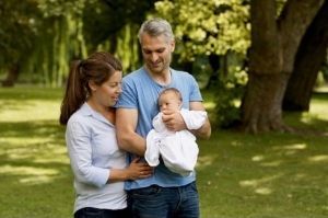 Ein Mann und eine Frau stehen in einem Park und schauen lächelnd auf einen Säugling, den der Mann auf dem Arm trägt.