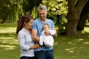 Ein Mann und eine Frau stehen in einem Park und schauen lächelnd auf einen Säugling, den der Mann auf dem Arm trägt. Ein Mann und eine Frau stehen in einem Park und schauen lächelnd auf einen Säugling, den der Mann auf dem Arm trägt.