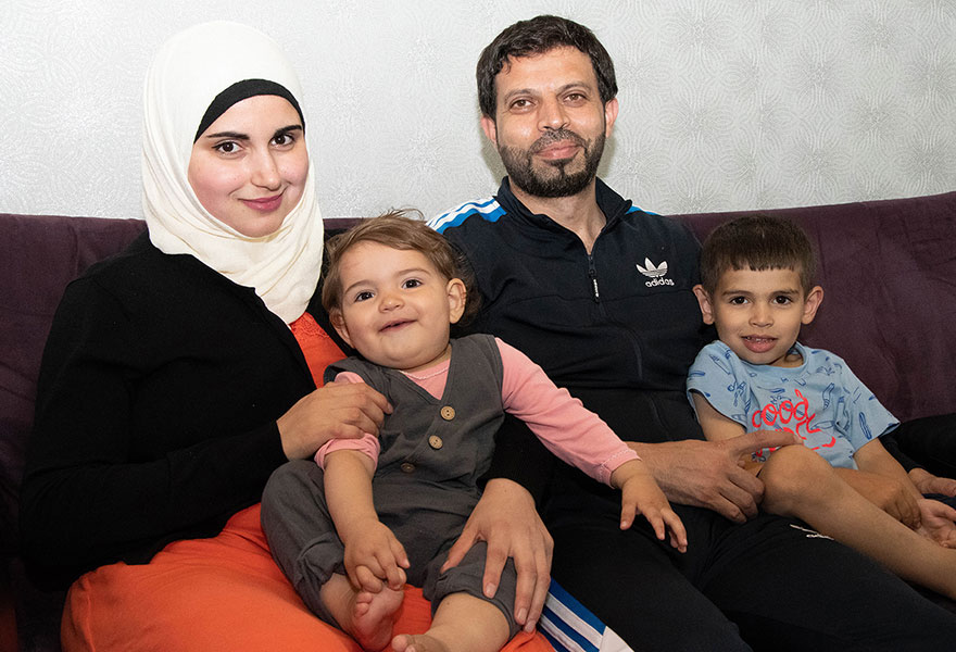 Das Ehepaar sitzt mit ihren Kindern auf dem Sofa. Alle lächeln in die Kamera.