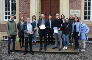 Zwölf Männer und Frauen stehen vor dem Eingang eines Gebäudes zum Gruppenbild positioniert auf einer Treppe. Zwei Männer in der Mittel halten Dokumente in den Händen. Zwölf Männer und Frauen stehen vor dem Eingang eines Gebäudes zum Gruppenbild positioniert auf einer Treppe. Zwei Männer in der Mittel halten Dokumente in den Händen.