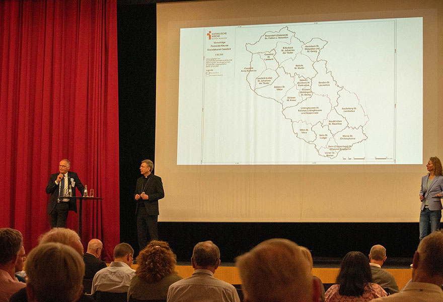 Auf einer Bühnen stehen Generalvikar Dr. Klaus Winterkamp und Weihbischof Dr. Stefan Zekorn. Im Hintergrund ist die Folie mit den pastoralen Räumen an eine Leinwand projiziert. Im Vordergrund sitzen Menschen.