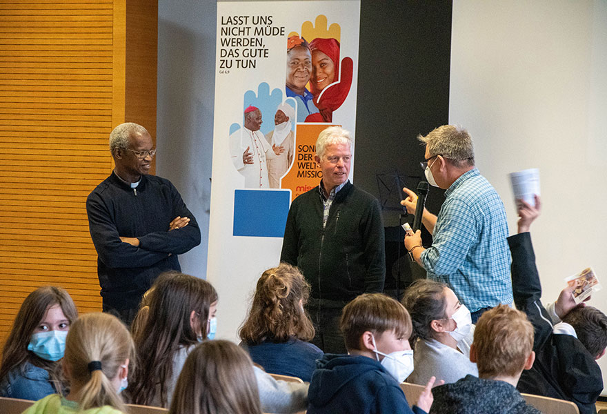 Im Vordergrund sind Schülerinnen und Schüler zu sehen. Dahinter sprechen Bischof Richard, Hans-Georg Hollenhorst und Thomas Verbücheln miteinander.