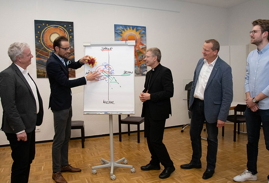 Auf dem Gruppenfoto schauen die Teilnehmer auf ein Flipchart, an dem Dr. Marius Stelzer etwas erläutert. 