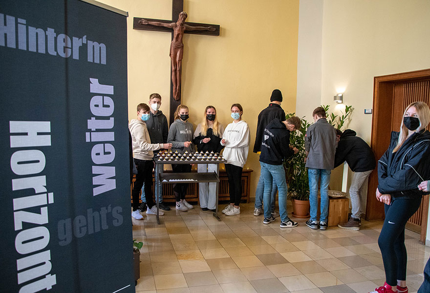 Im Vordergrund steht ein Aufsteller mit der Aufschrift "Hinterm Horizont gehts weiter". Dahinter stellen unter einem Kreuz Schülerinnen und Schüler LED-Teelichter auf.