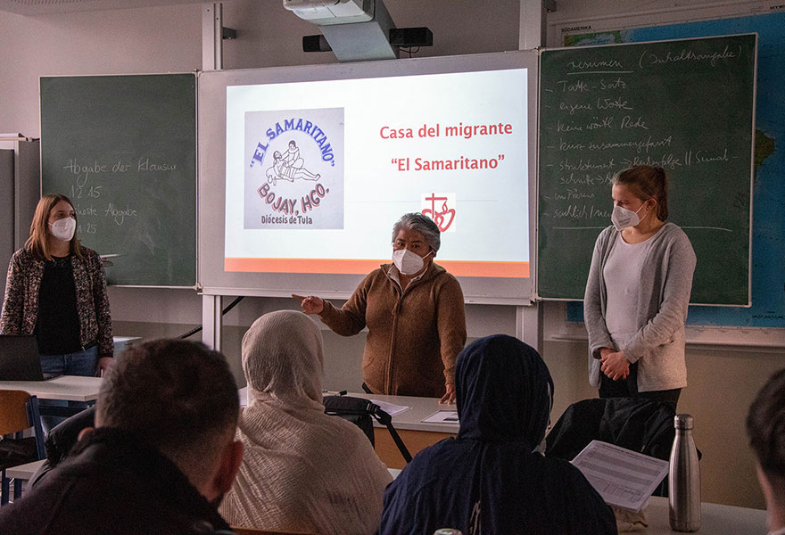 Drei Frauen stehen vor einer Klasse. Im Hintergrund ist eine Präsentation über das Migrantenhaus an der Tafel zu sehen.