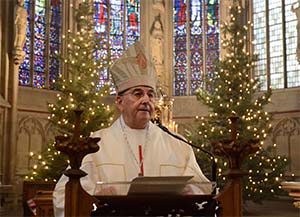 Bischof Dr. Felix Genn steht am Abo der St.-Lamberti-Kirche Münster. Im Hintergrund stehen im Altarraum große, mit Kerzen geschmückte Tannenbäume.