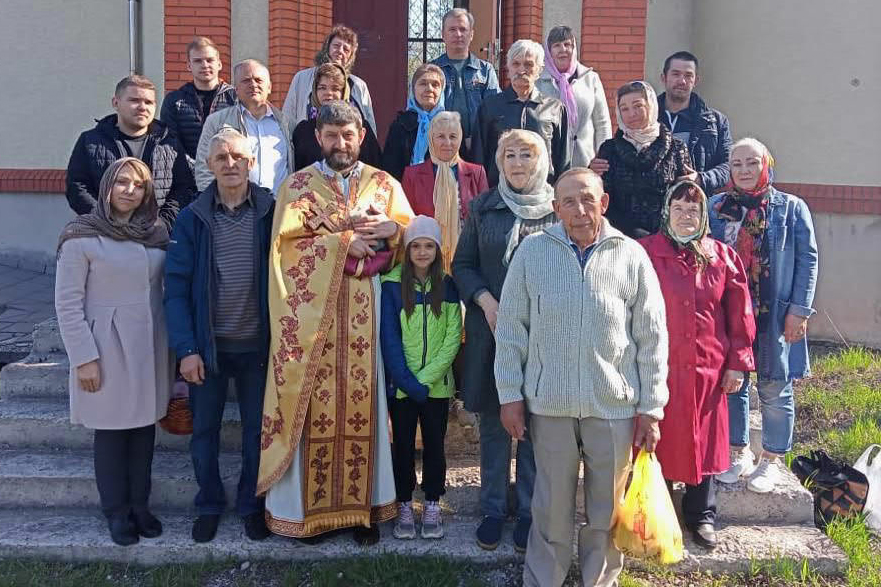 Ein Priester, Kinder und Erwachsene stehen lächelnd auf den Stufen einer Kirchentür. 