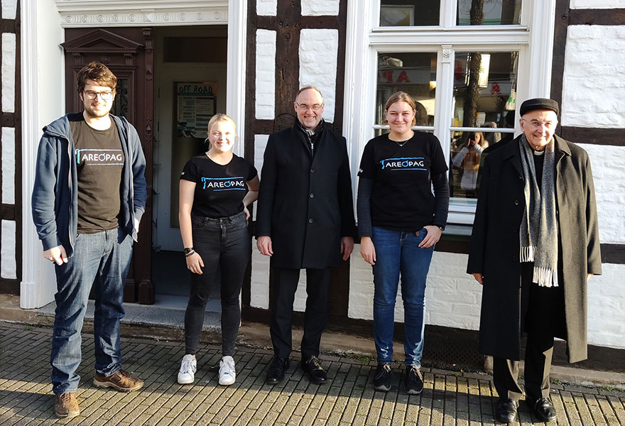 Die Mitarbeitenden des Areopags haben sich mit Weihbischof Lohmann und Bischof Genn zum Gruppenfoto vor dem Gebäude aufgestellt.