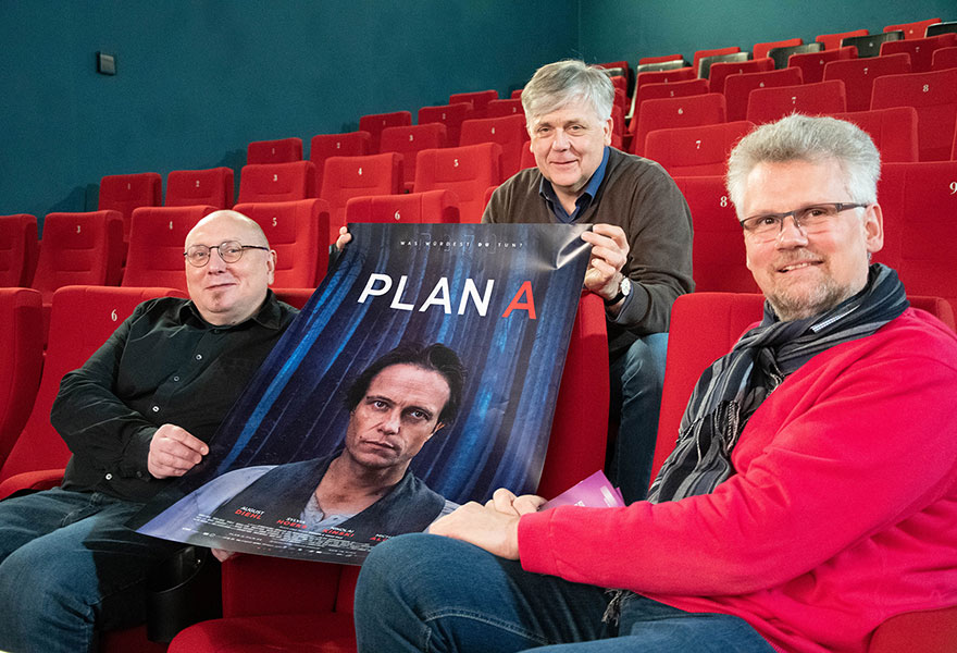 Drei Männer sitzen in Kinosesseln und halten ein Plakat in der Hand.