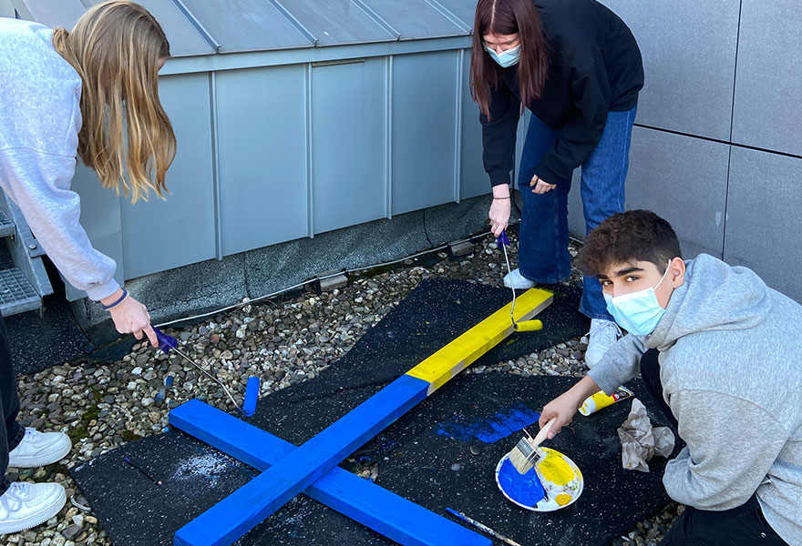 Zwei Schülerinnen und ein Schüler streichen ein großes Kreuz in blau und gelb.