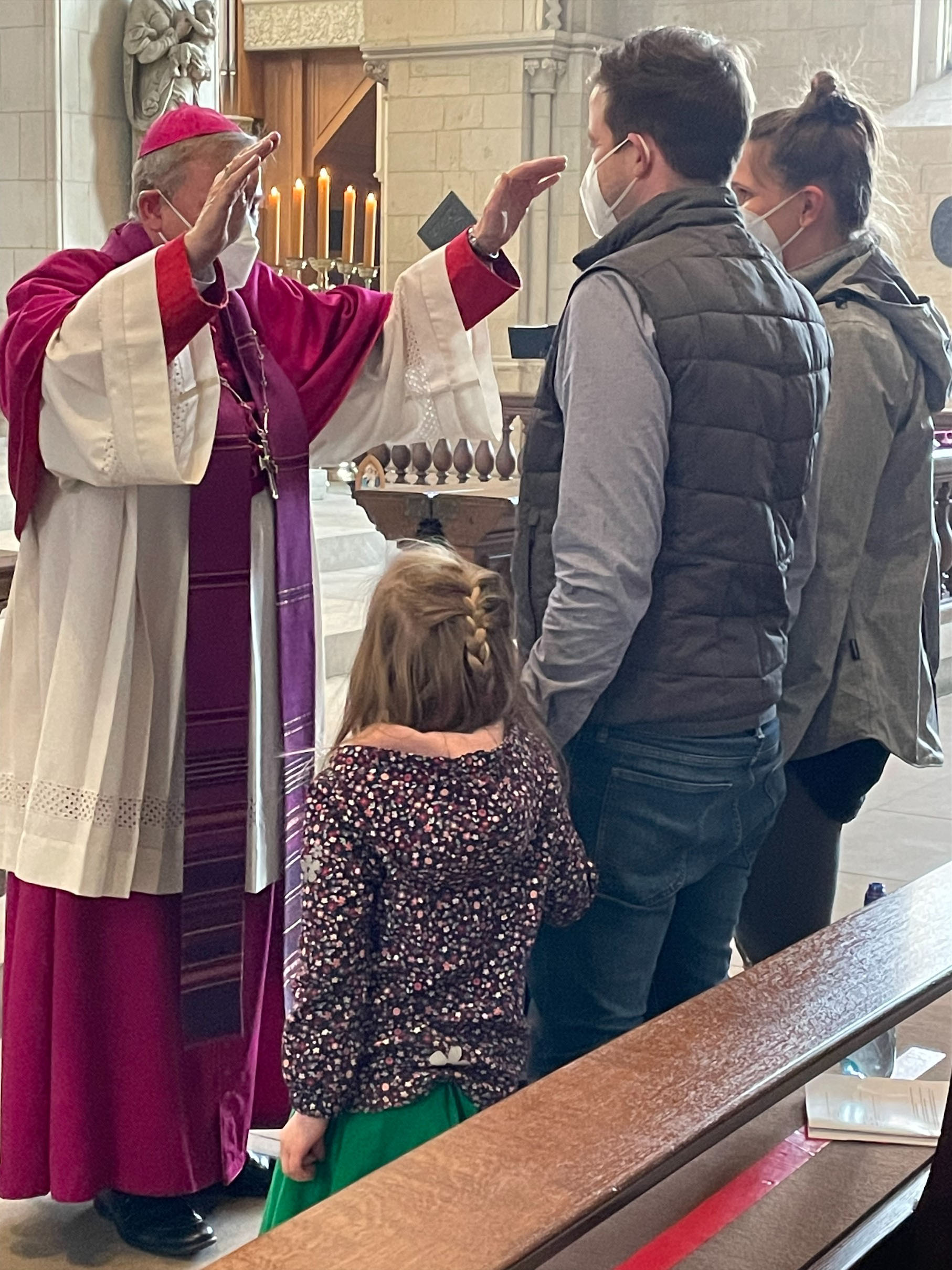 Der Weihbischof, links im Bild stehend, erhebt segnend die Arme über einer Frau und einem Mann, die mit ihrer kleinen Tochter im Dom vor ihm stehen. 