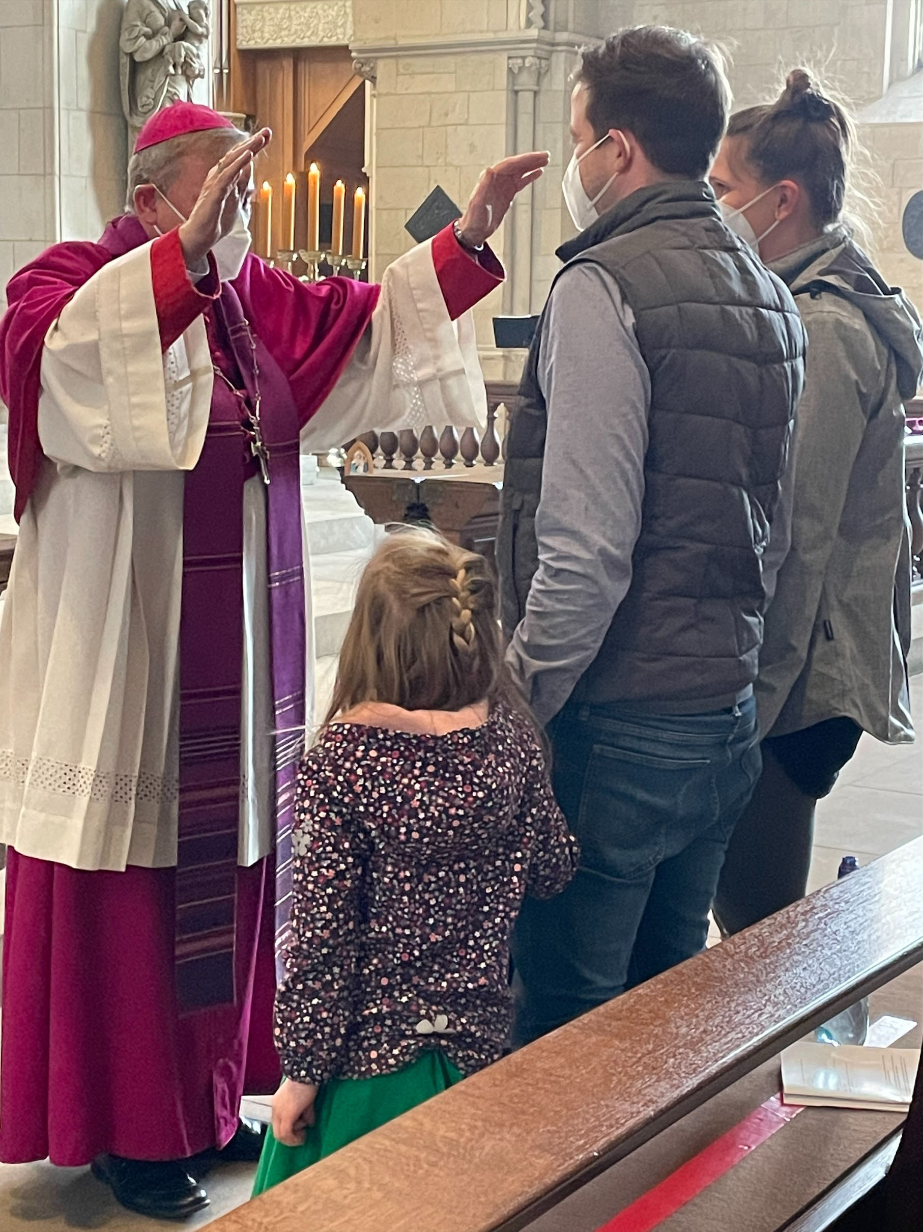 Der Weihbischof, links im Bild stehend, erhebt segnend die Arme über einer Frau und einem Mann, die mit ihrer kleinen Tochter im Dom vor ihm stehen. Der Weihbischof, links im Bild stehend, erhebt segnend die Arme über einer Frau und einem Mann, die mit ihrer kleinen Tochter im Dom vor ihm stehen.