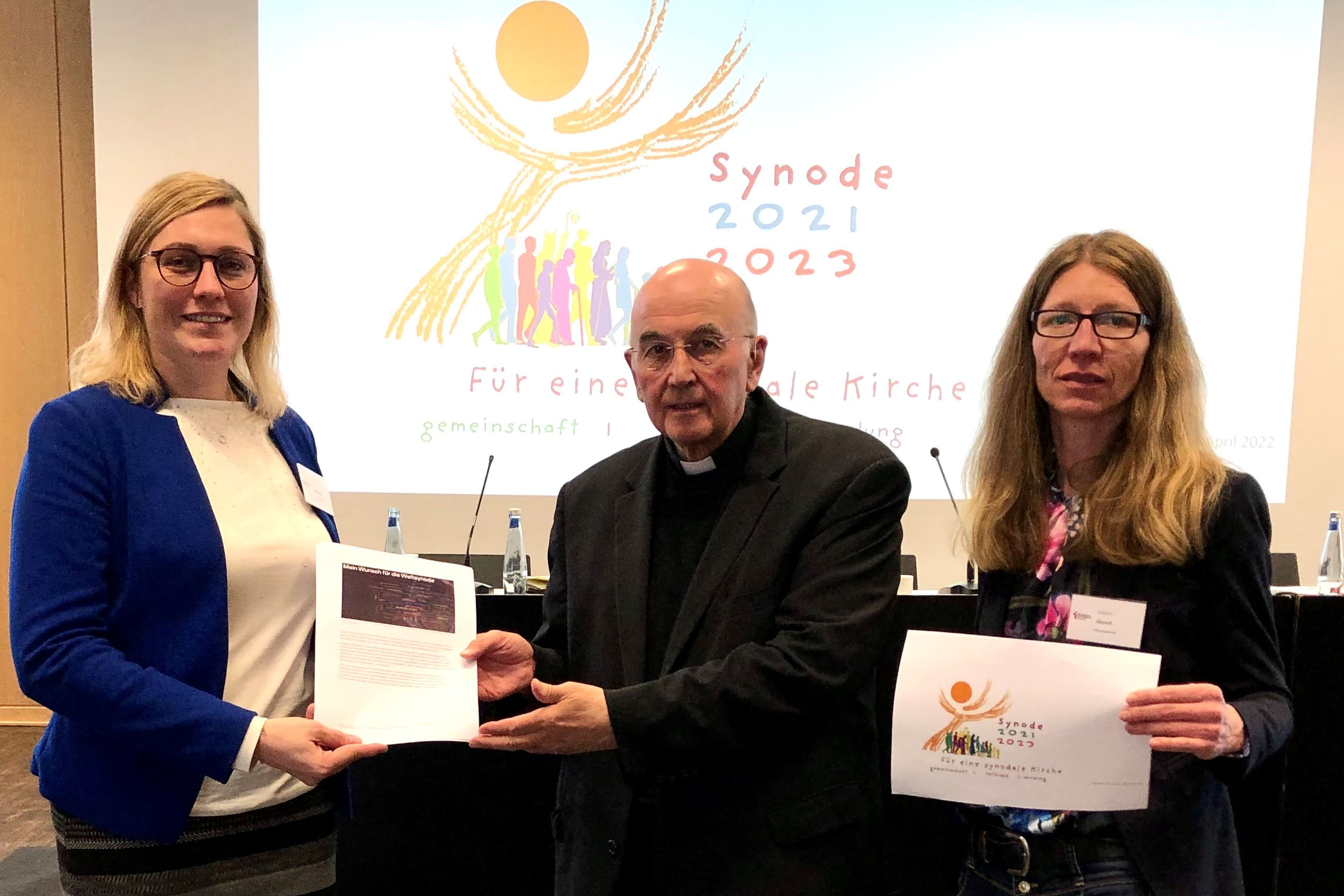 Kerstin Stegemann (links) und Susanne Deusch übergeben die Rückmeldungen in Papierform an Bischof Felix Genn. Im Hintergrund ist das Logo der Weltsynode zu erkennen.