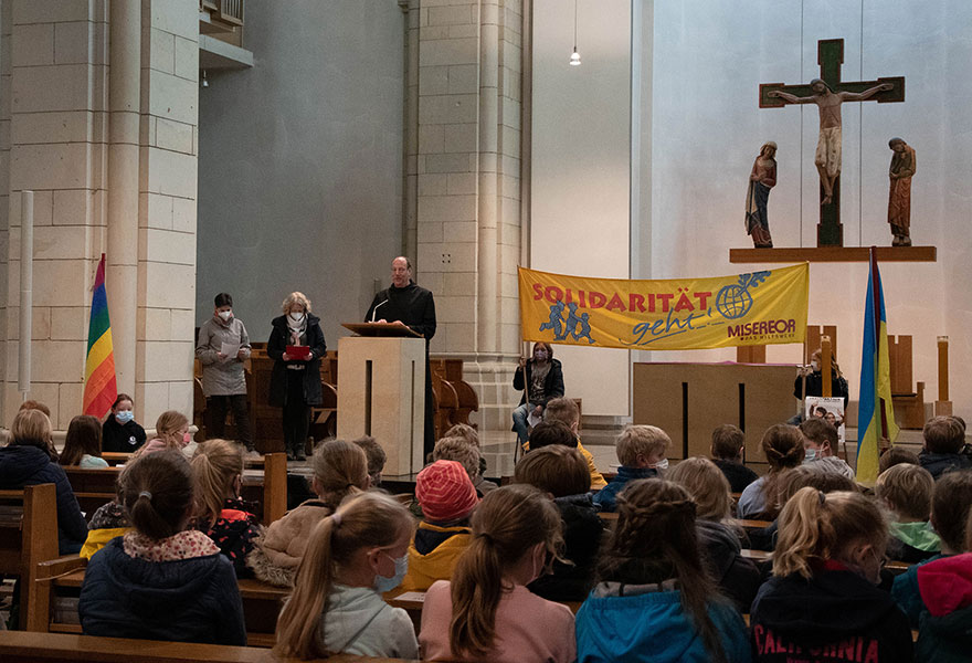 In der Klosterkirche sitzen vorne viele Schülerinnen und Schüler, die von Pater Norbert am Ambo begrüßt werden.