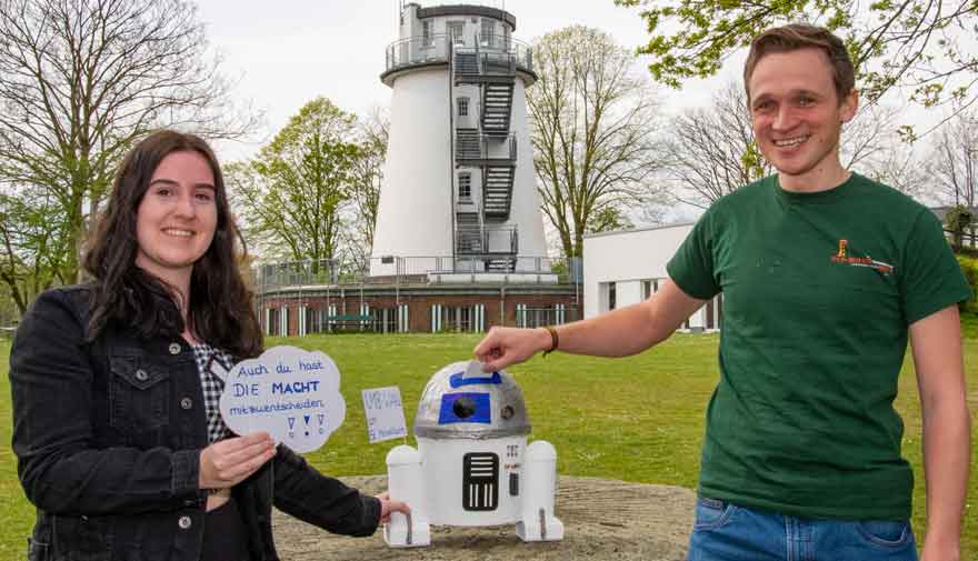 Eine Frau und ein Mann stehen vor einem Turm, in der Mitte steht ein gebastelter Roboter.