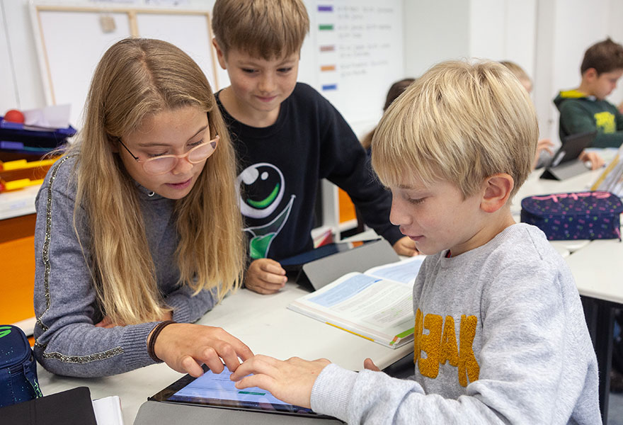 Drei Kinder sitzen im Klassenraum und schauen sich etwas auf einem Tablet an.