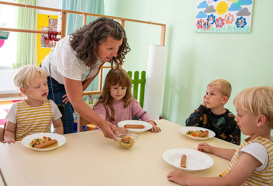 Eine Frau stellt vier Kindern ein Schälchen mit Eintopf auf den Tisch.