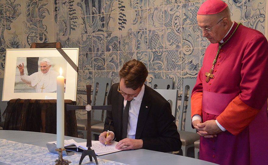 Links ein Foto des verstorbenen Benedikt XVI. mit Trauerschleife, in der Mitter trägt sich Ministerpräsident Wüst in das auf einem Tisch liegende Kondolenzbuch ein. Rechts daneben steht Bischof Dr. Felix Genn.
