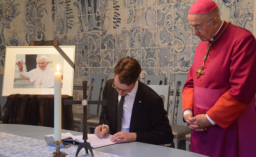 Links ein Foto des verstorbenen Benedikt XVI. mit Trauerschleife, in der Mitter trägt sich Ministerpräsident Wüst in das auf einem Tisch liegende Kondolenzbuch ein. Rechts daneben steht Bischof Dr. Felix Genn. Links ein Foto des verstorbenen Benedikt XVI. mit Trauerschleife, in der Mitter trägt sich Ministerpräsident Wüst in das auf einem Tisch liegende Kondolenzbuch ein. Rechts daneben steht Bischof Dr. Felix Genn.