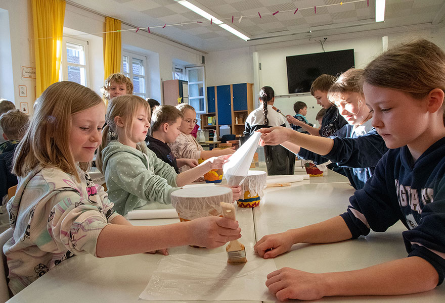 Kinder sitzen am Tisch und kleben Pergamentpapier auf die angemalte Trommel.