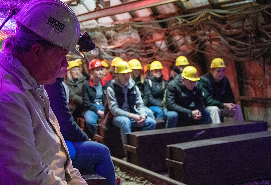 Ein Mann mit weißem Anzug und Helm sitzt im Vordergrund, dahinter sieht man Jugendliche, die gelbe Helme tragen, in einem Schacht.