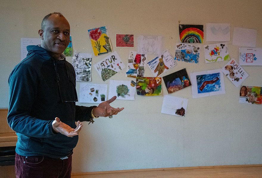 Im Vordergrund steht der nigerianische Künstler und spricht. Hinter ihm hängen verschiedene Bilder an der Wand. Im Vordergrund steht der nigerianische Künstler und spricht. Hinter ihm hängen verschiedene Bilder an der Wand.