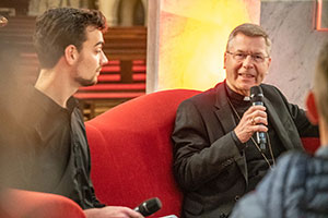 Der Moderator und der Weihbischof sitzen auf einer roten Couch.