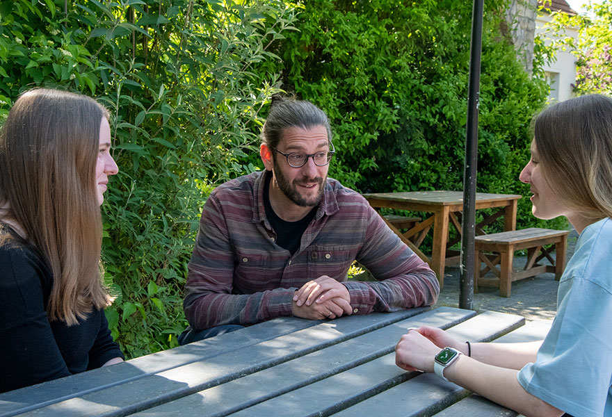 Amelie Liesen und Lara Cord-to-Krax sitzen gemeinsam mit Matthias Kortmann an einem Tisch und sprechen miteinander.