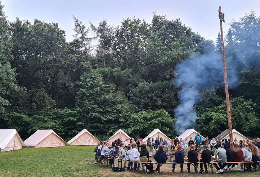 Kinder und Jugendliche sitzen um ein Lagerfeuer herum. Dahinter sieht man zahlreiche Zelte.