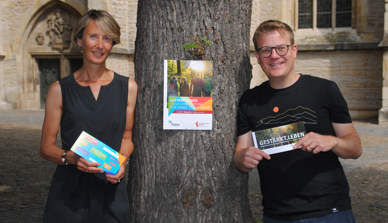 Stefanie Uphues und Christoph Aperdannier lehnen sich an einen großen Baumstamm. In den Händen halten Sie den Flyer mit Informationen über das Angebot zur Firmvorbereitung.