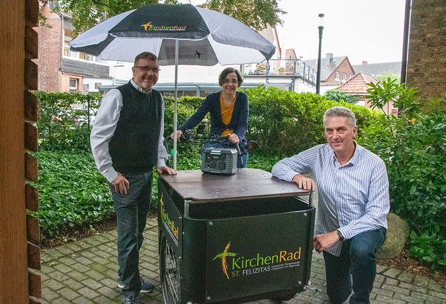 Links und rechts neben der Transportbox des Rades stehen zwei Männer, in der Mitte sitzt eine Frau auf dem Sattel und hält den Lenker.