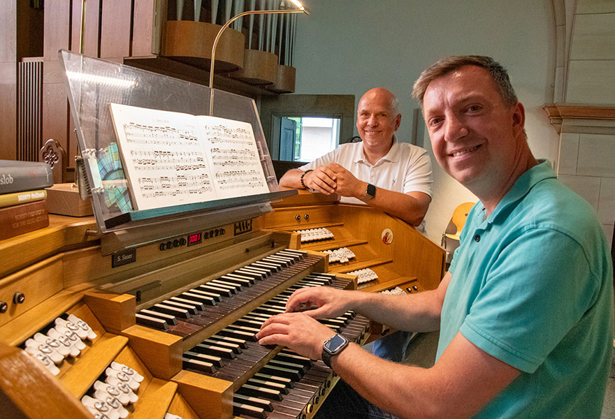 Björn Buchholz sitzt an der Orgel und lächelt in die Kamera. Im Hintergrund steht sein Lehrer Detlef Steinbrenner.
