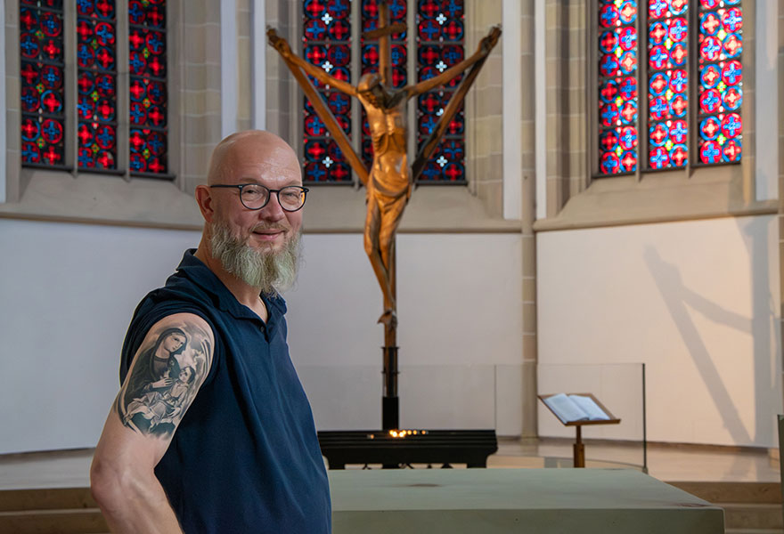 Matthias Laarmann steht vor dem Coesfelder Kreuz in der Kirche und zeigt sein Oberarmtattoo mit der Darstellung der Muttergottes mit Kind.