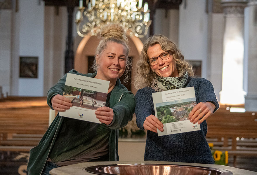 Zwei Frauen stehen am Taufbecken in der Kirche und halten den Kinderkirchenführer in die Kamera.