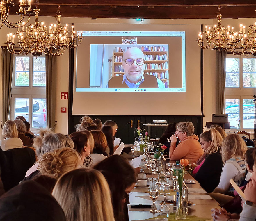 Menschen in einem Tagungsaal blicken auf eine Leinwand. Auf dieser Leinwand ist Mann zu sehen, im Hintergrund ist ein Bücherregal zu erkennen.