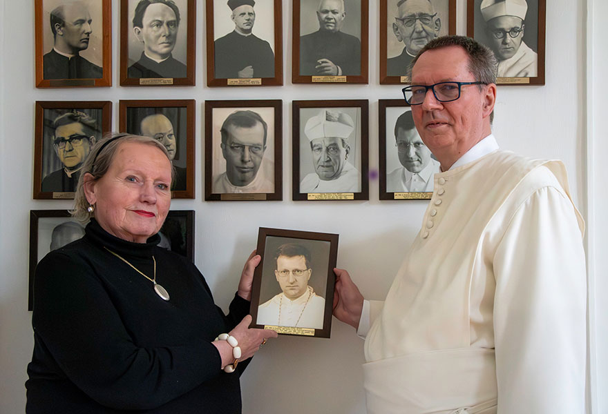 Monika Löchter und Pater Dominik Kitta stehten vor einer Wand, an der viele Fotos aufgehängt sind.