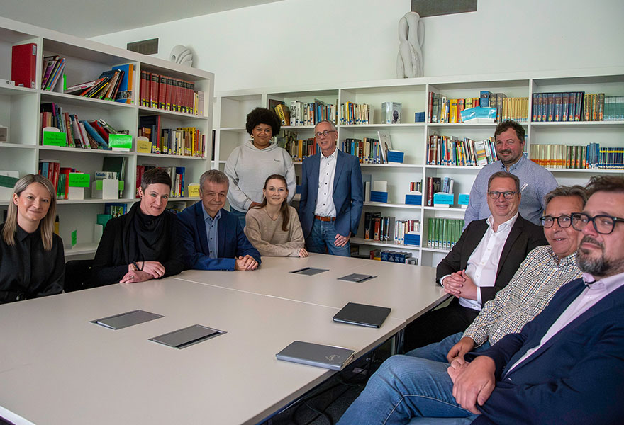 Die Beteiligten sitzen rund um einen Tisch vor Bücherregalen in der Bibliothek.