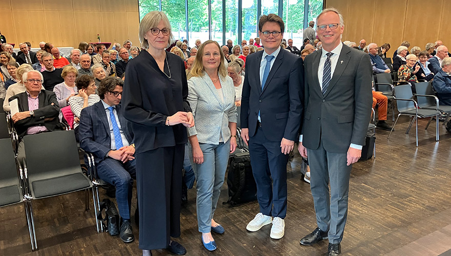 Ein großer Saal in dem viele sitzende Menschen zu sehen sind. Davor stehen (von links) Maria Kröger, Katharina Klöcker, Philipp Lenz und Klaus Winterkamp und schauen in die Kamera.