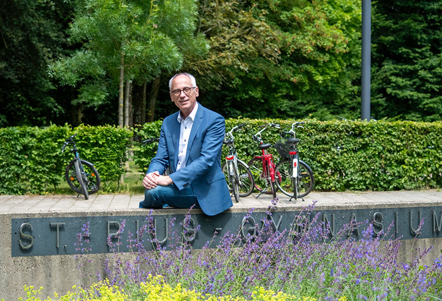 Dr. Norbert Just sitzt auf einer Mauer, auf der der Schriftzug St.-Pius-Gymnasium steht. Dr. Norbert Just sitzt auf einer Mauer, auf der der Schriftzug St.-Pius-Gymnasium steht.