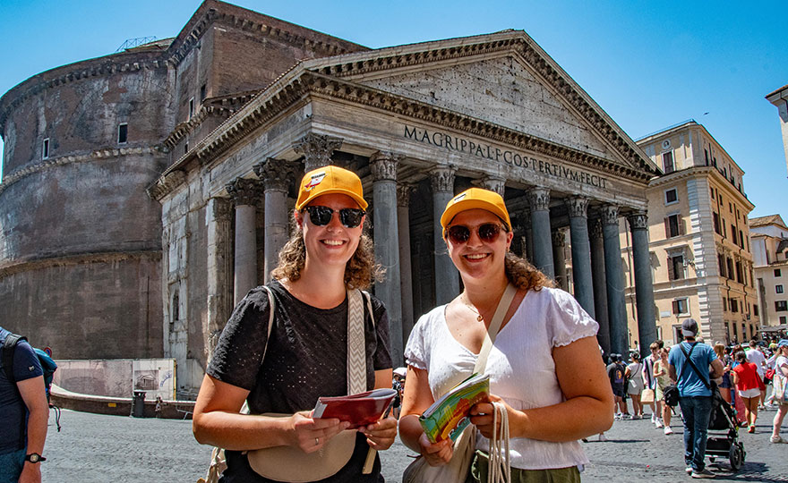 Zwei junge Frauen mit gelben Baseball-Caps und Sonnenbrille stehen vor einer Sehenswürdigkeit in Rom und lächeln in die Kamera.