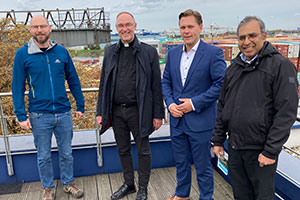 Vier Männer stehen auf einem Balkon. Im Hintergrund ist Teile des Duisburger Hafens sichtbar.