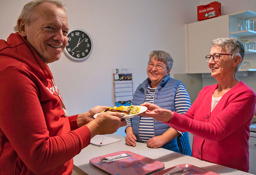 Eine Frau überreicht einem Mann einen Teller mit Essen. Alle schauen freundlich.