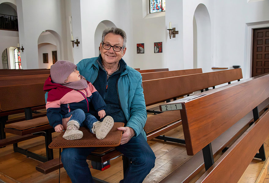 Herr Hülsken sitzt in einer Kirchenbank, seine Enkelin sitzt auf einem Kissen auf seinen Beinen.
