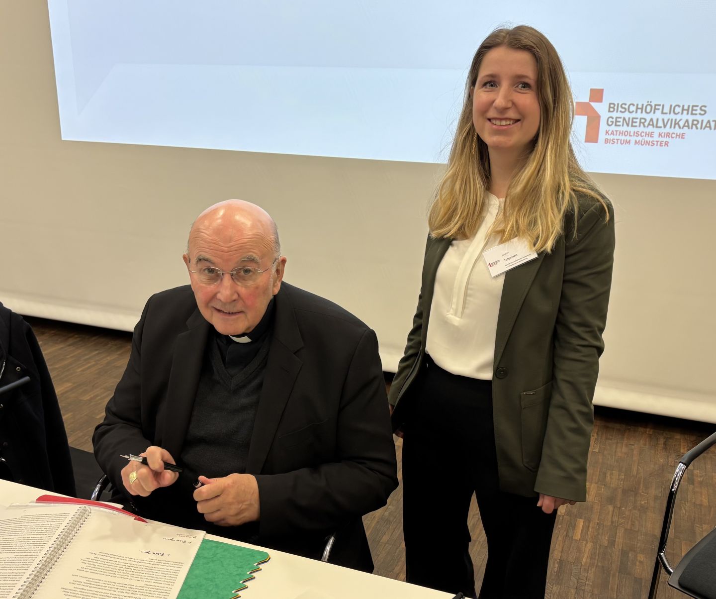 Bischof Dr. Felix Genn sitzt an einem Tisch und hält eine Stift in der Hand, auf dem Tisch liegt eine Mappe mit Dokumenten. Neben ihm steht Jasmin Telgmann.