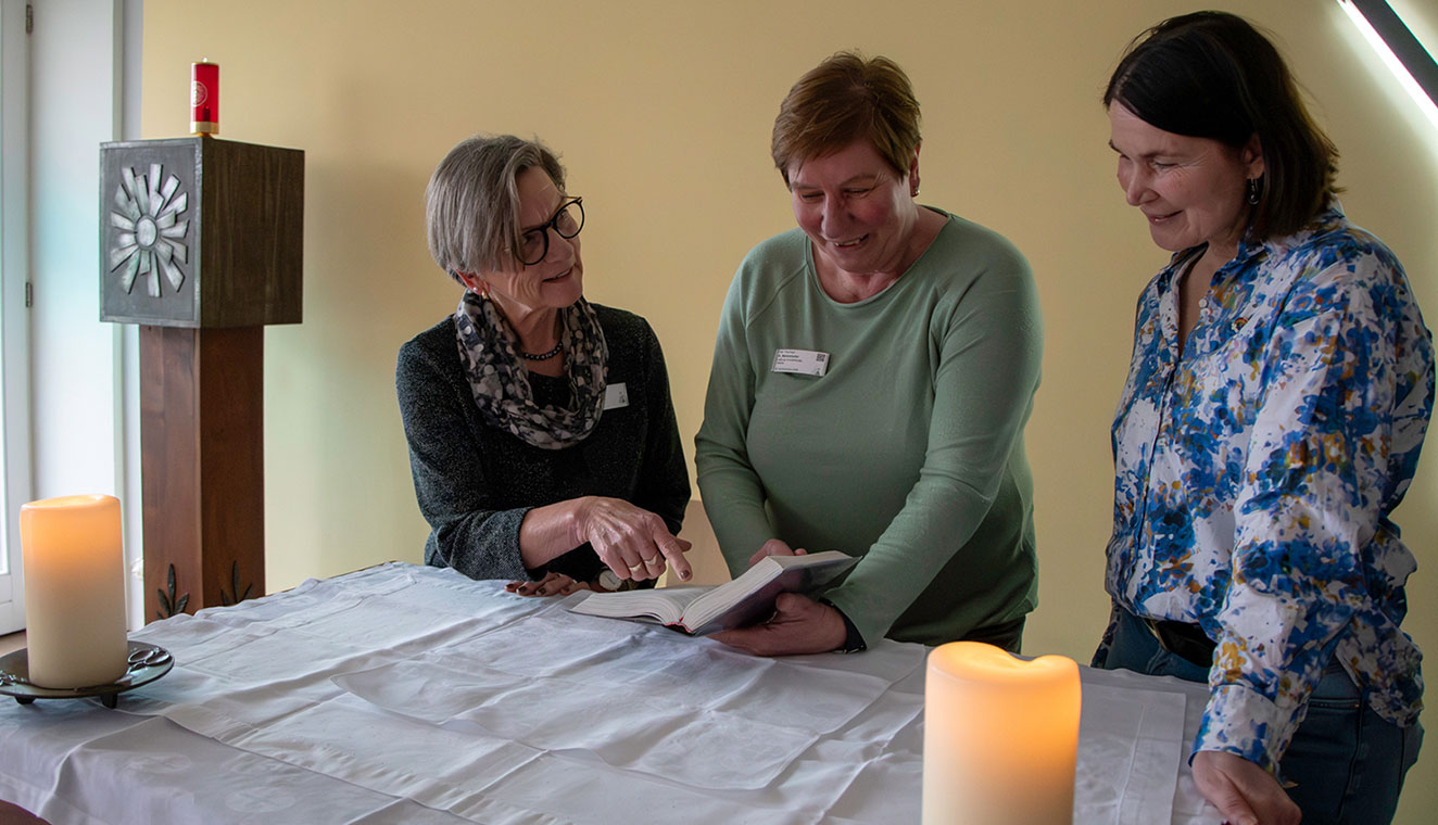 Drei Frauen stehen hinter einem mit Kerzen ausgestatteten Hausaltar und blättern in einem Buch. Im Hiuntergrund ist ein tabernakel zu sehen. 