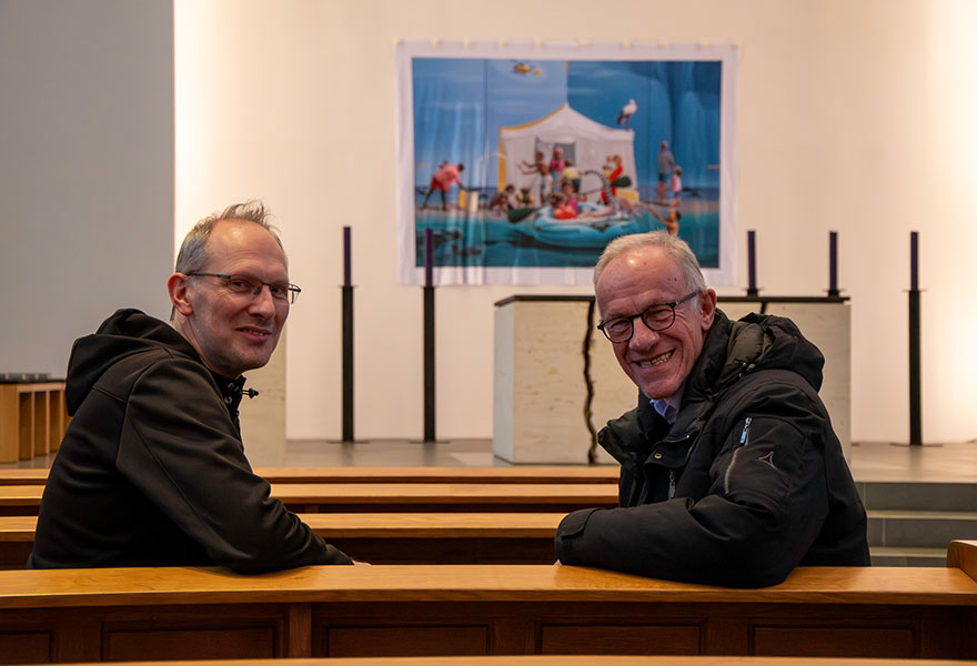 Ralf Blasi und Johannes Hammans sitzen in einer Kirchenbank und lächeln in die Kamera.