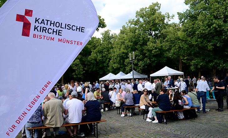 Auf dem Domplatz sitzen Menschen an Biertischen, im Hintergrund sind Pavillons aufgestellt. Im Vordergrund sieht man eine Werbefahne mit dem Logo des Bistums Münster.