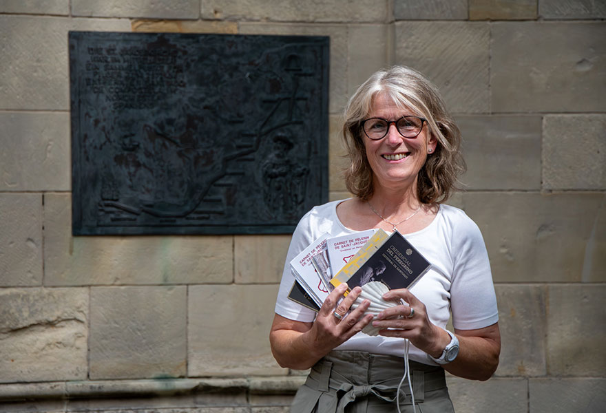 Ulla Kortüm steht an einer Bronzetafel und hat die Jakobsmuschel und einige Pilgerbücher in der Hand.